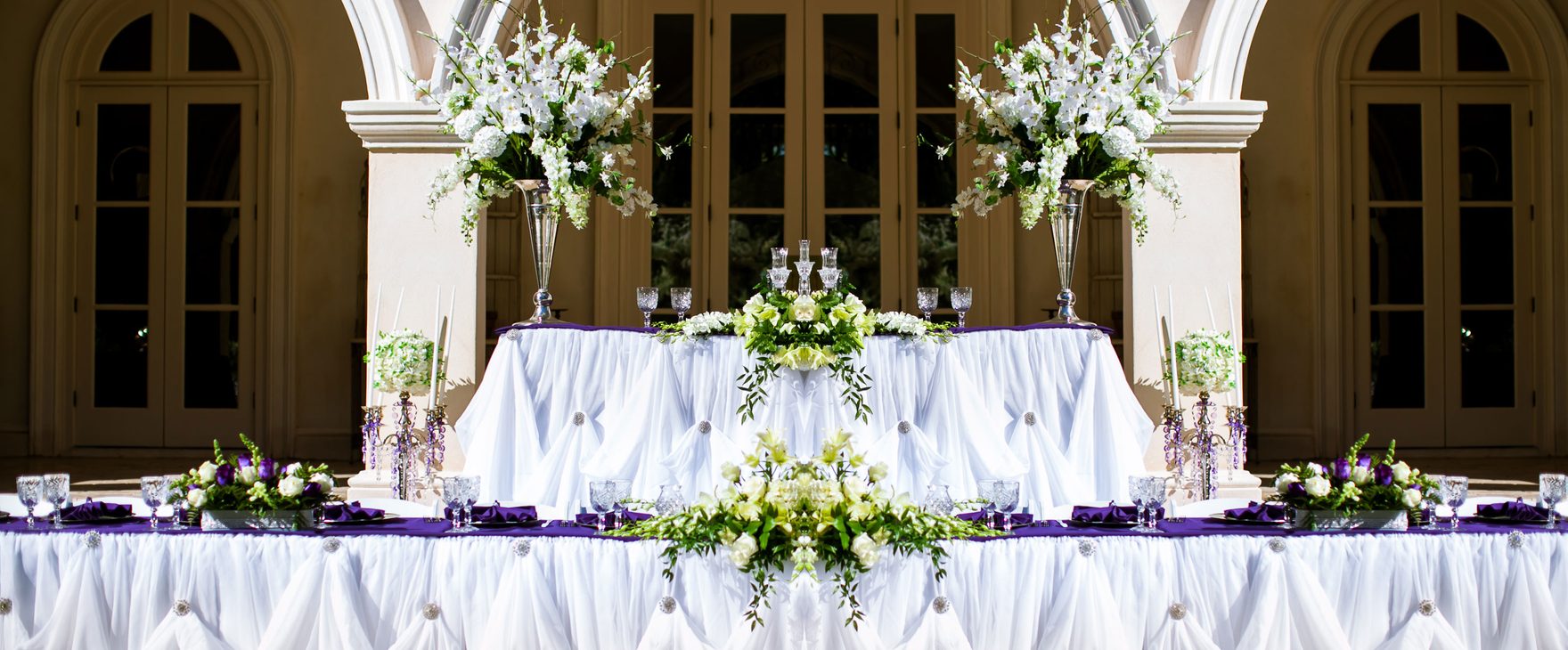 Head table draped in white with tall floral arrangements beneath stone arches