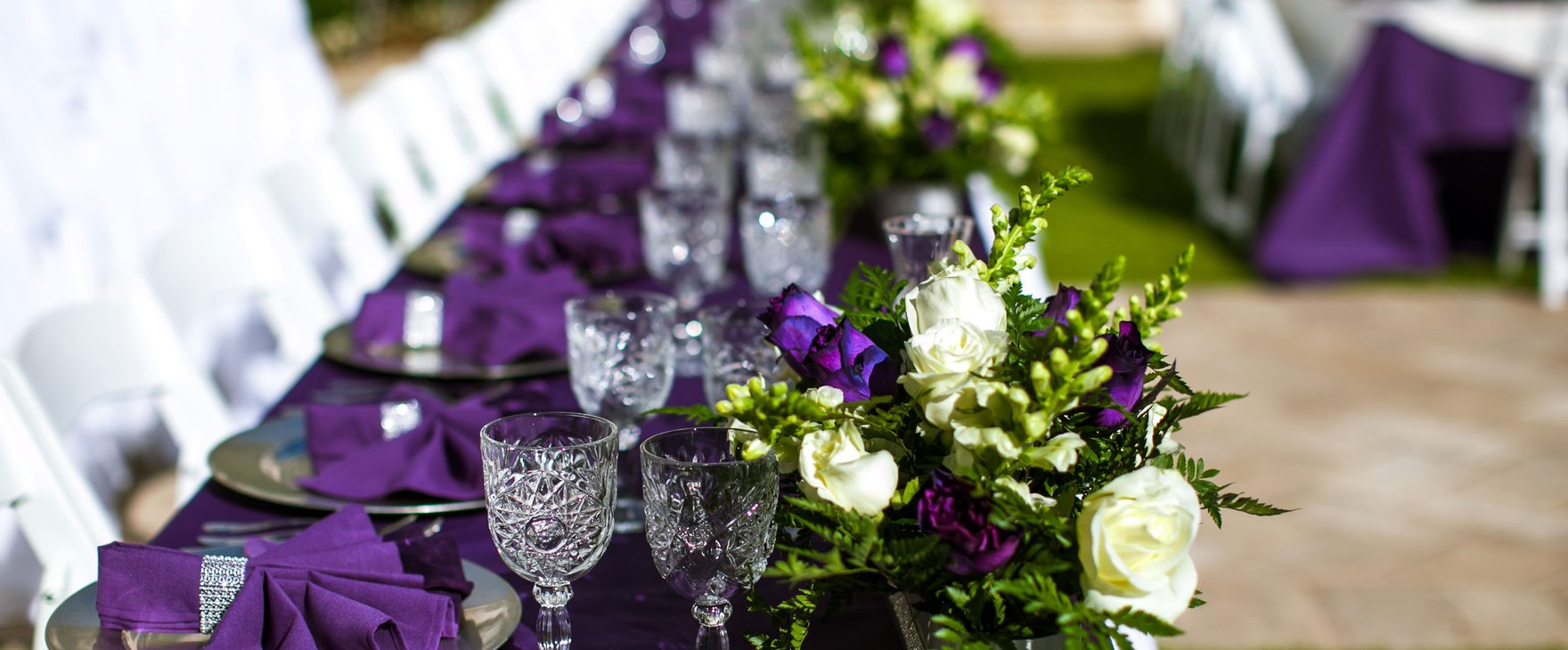Crystal glassware, purple napkins and white roses along a banquet table