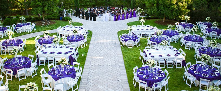 Garden reception of round tables with purple linens around a stone aisle