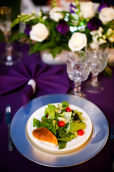 Plated salad beside a centerpiece of white roses and purple flowers